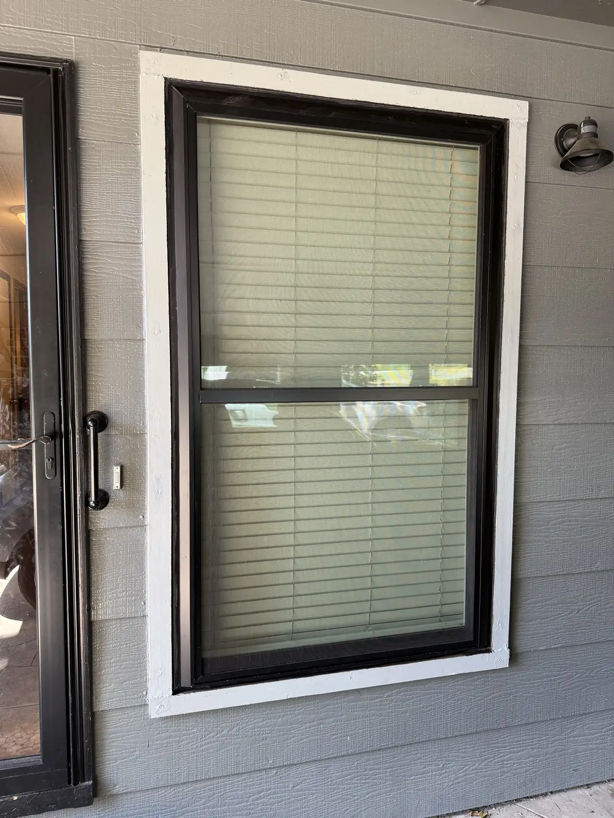 New black-frame double-hung window with white trim on gray siding beside a patio door